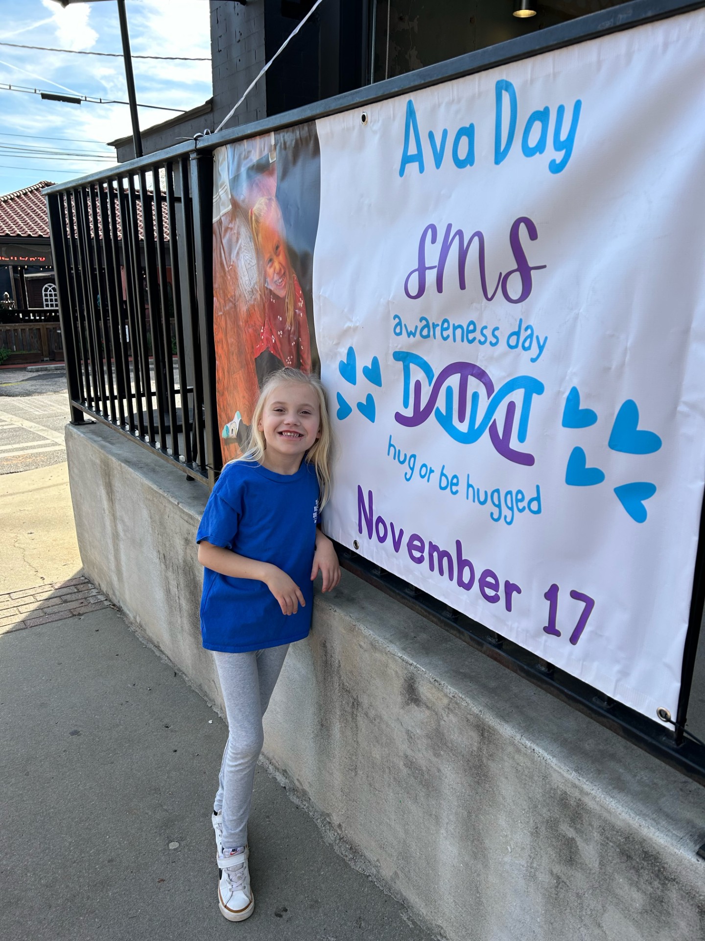 A blonde girl in a blue t-shirt stands next to a sign proclaiming November 17th as Ava Day