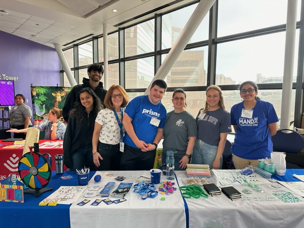 A group smiles and poses behind a PRISMS booth at a conference. The table is full of brochures and PRISMS branded merch