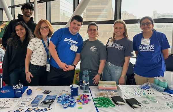 Awareness-Clinic A group smiles and poses behind a PRISMS booth at a conference. The table is full of brochures and PRISMS branded merch